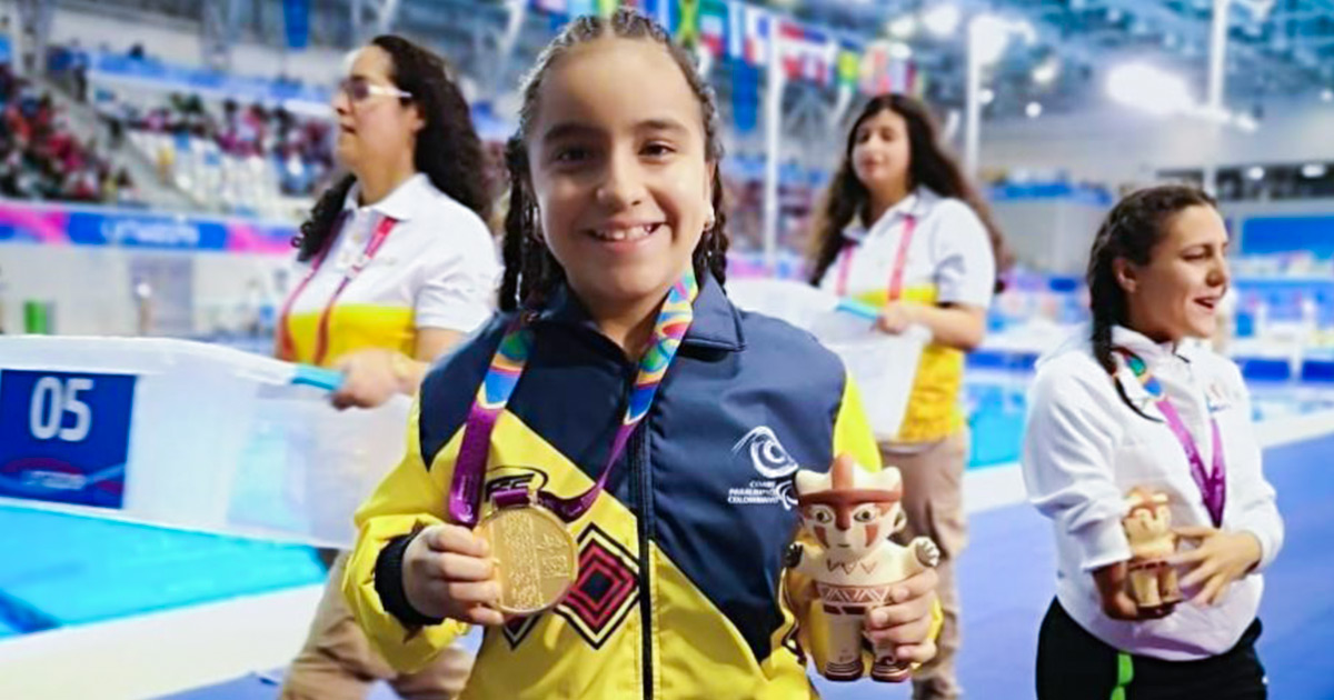 Para nadadora Sara Vargas posa sonriente a cámara sosteniendo su medalla de oro en una mano y su Cuchimilco en la otra. 