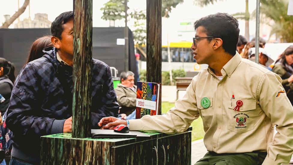 Hombre junto a voluntario de Sernanp para recibir información sobre la herramienta de huella de carbono, en los Juegos Lima 2019. 